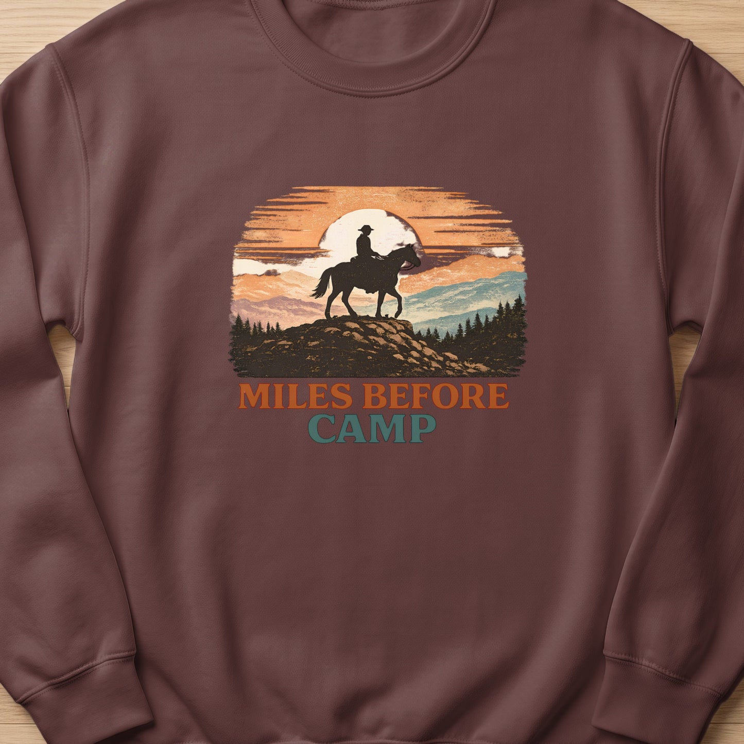 Crewneck sweatshirt with a sunset graphic of a cowboy on horseback with text "MILES BEFORE CAMP".