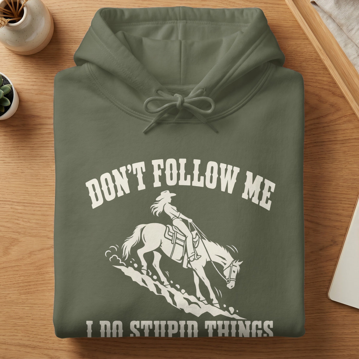 A folded olive-green hoodie with white text reading DON'T FOLLOW ME I DO STUPID THINGS and an illustration of a cowgirl riding a horse.