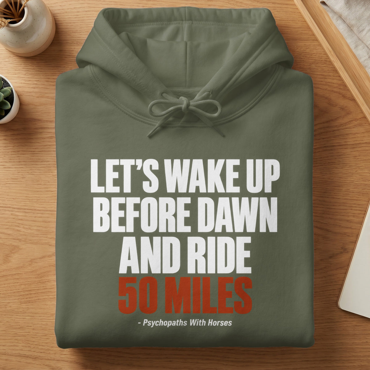 An olive green hoodie folded on a wooden table with white and red text.