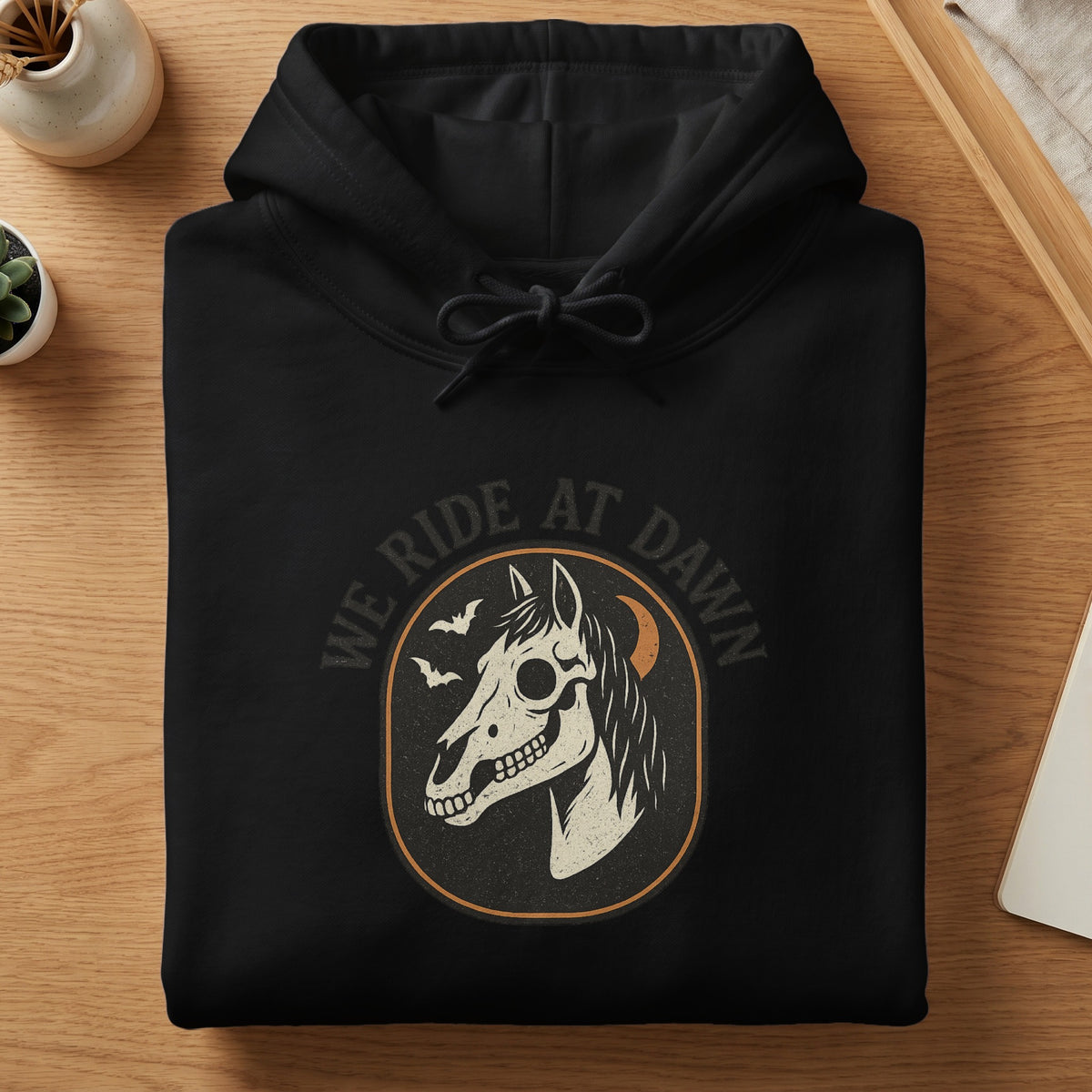Folded black hoodie featuring a skeleton horse graphic and the text We Ride At Dawn.