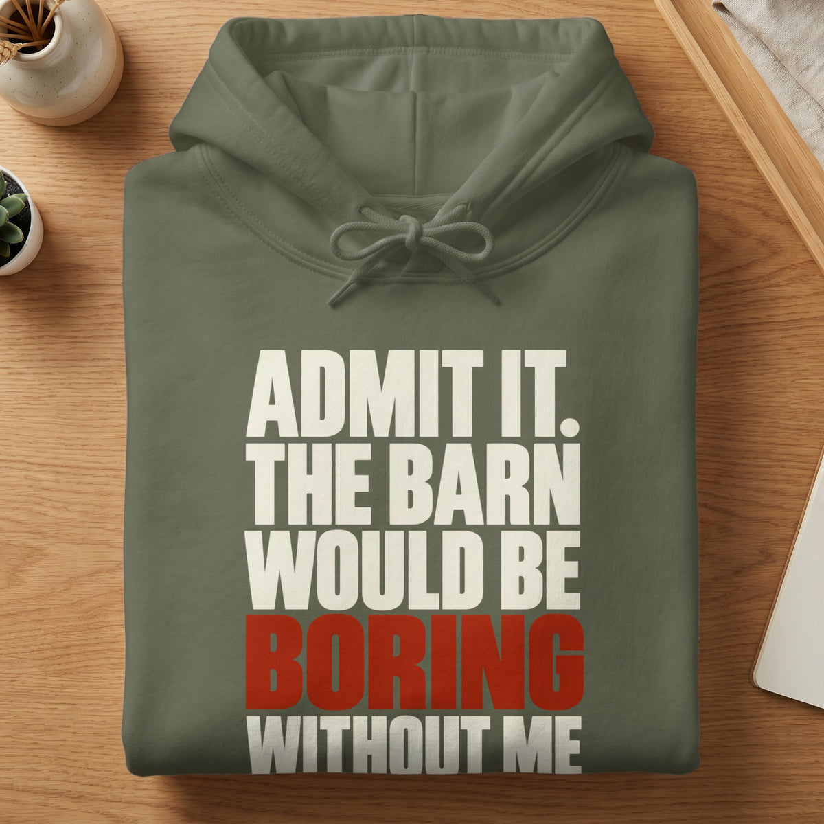 An olive green hoodie folded on a wooden surface with white and red text reading ADMIT IT. THE BARN WOULD BE BORING WITHOUT ME.