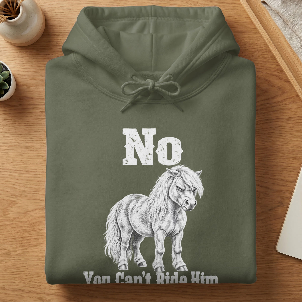 A folded olive green hoodie featuring white text reading No You Can't Ride Him alongside a sketched pony graphic.
