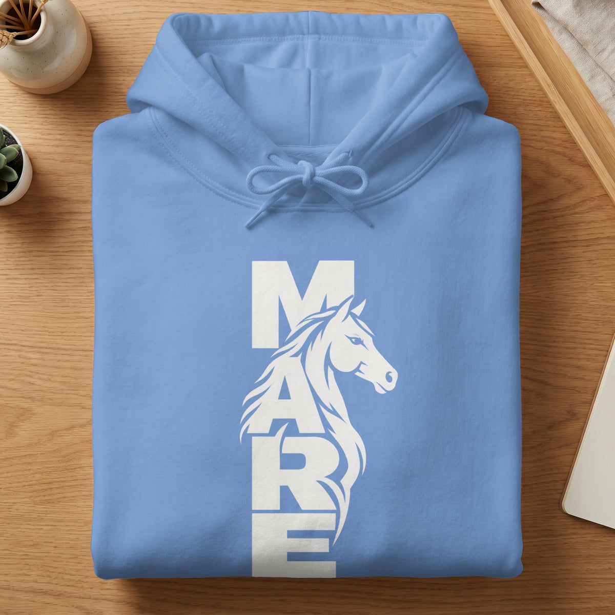 Folded light blue hoodie with white vertical text reading MARE and a horse graphic on a wooden table.