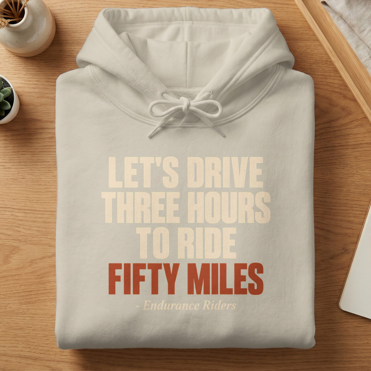 A folded beige hoodie with the text Let's Drive Three Hours to Ride Fifty Miles on a wooden table.