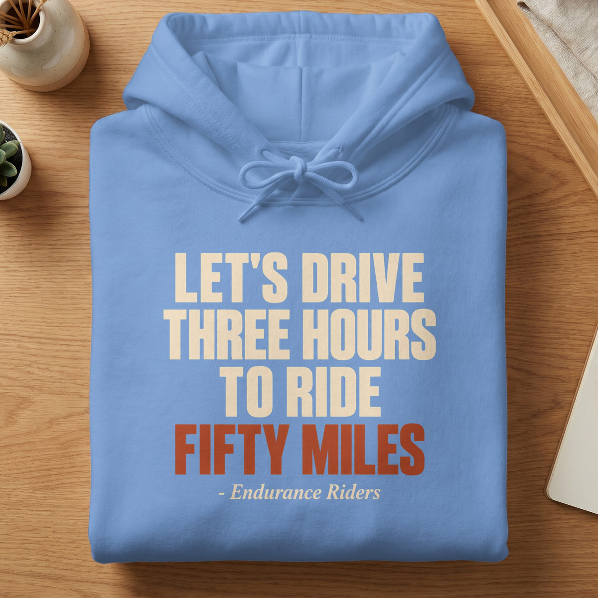 A folded light blue hoodie with white and orange text that reads, Let's Drive Three Hours To Ride Fifty Miles - Endurance Riders.