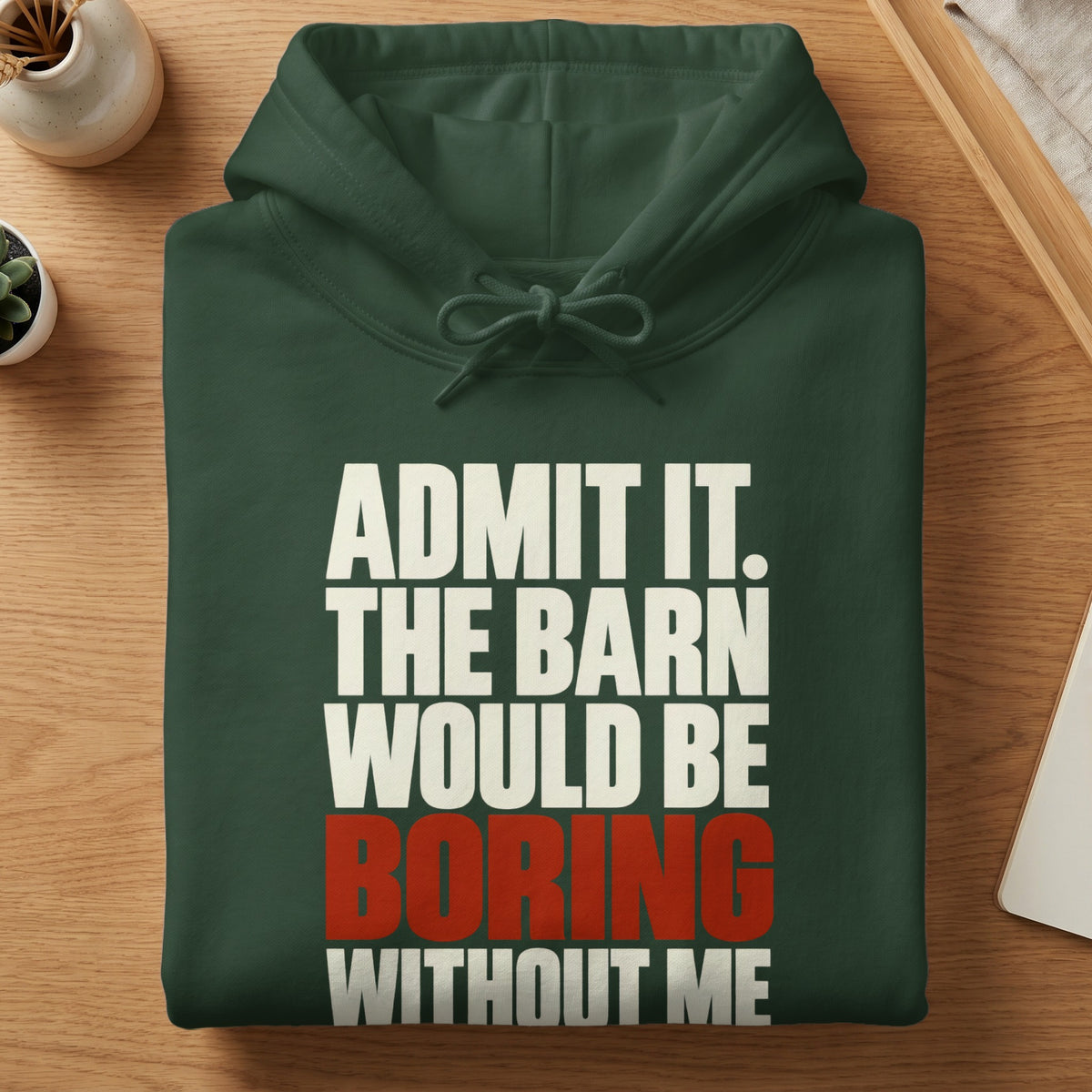 A neatly folded dark green hoodie with white and red text reading ADMIT IT. THE BARN WOULD BE BORING WITHOUT ME.