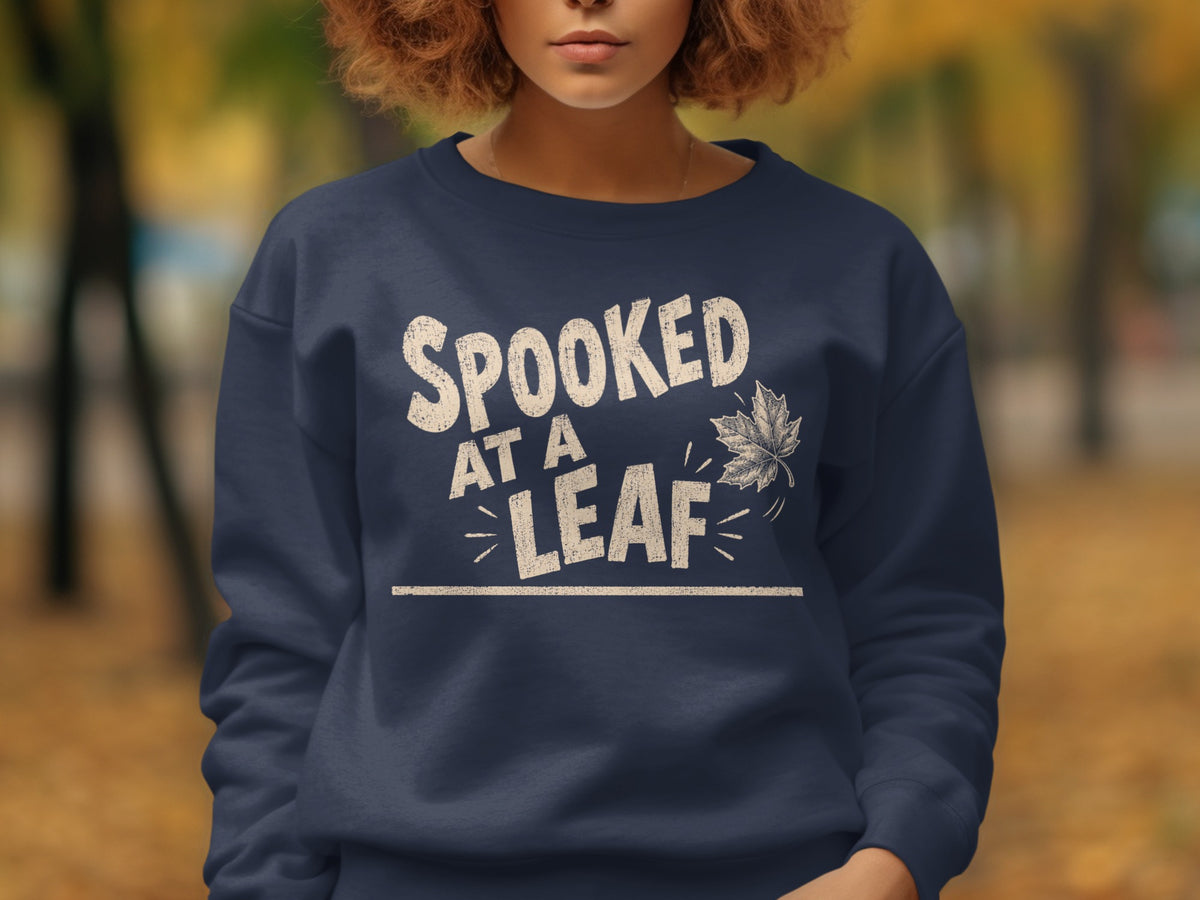 A woman wearing a dark blue crewneck sweatshirt with the white text Spooked At A Leaf and a falling maple leaf graphic.