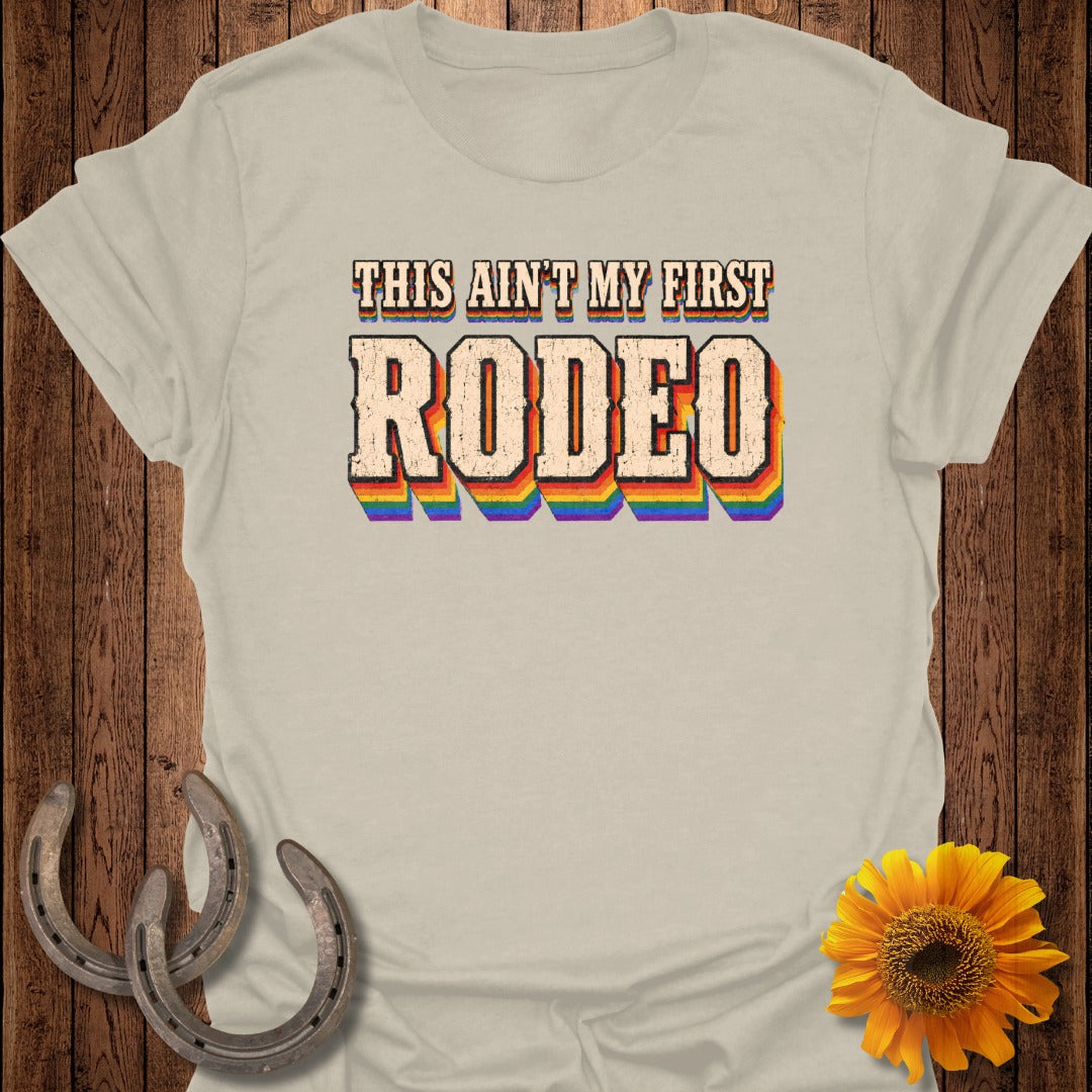 Cream t-shirt with "THIS AIN'T MY FIRST RODEO" in rainbow lettering.