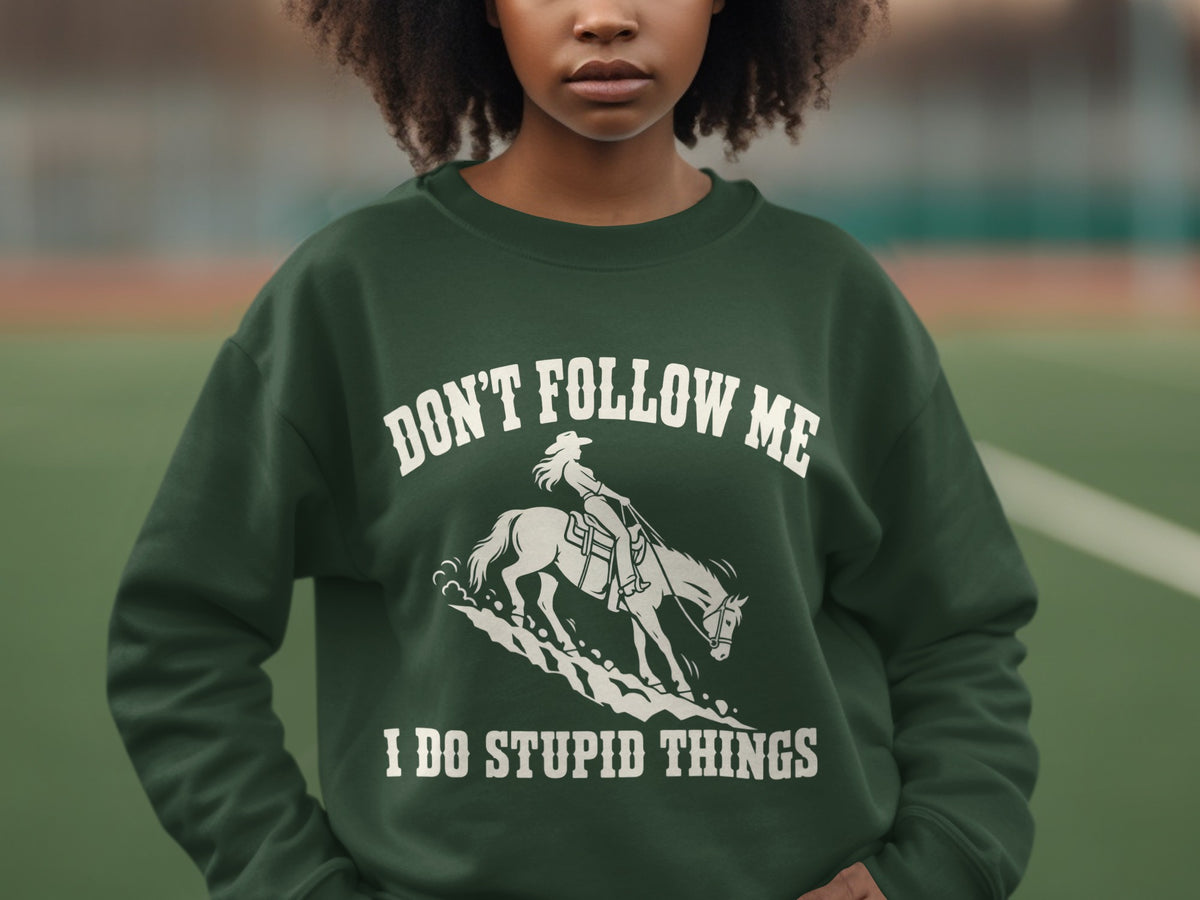 A person wears a dark green sweatshirt with white text reading "DON'T FOLLOW ME I DO STUPID THINGS" and a graphic of a person riding a horse.