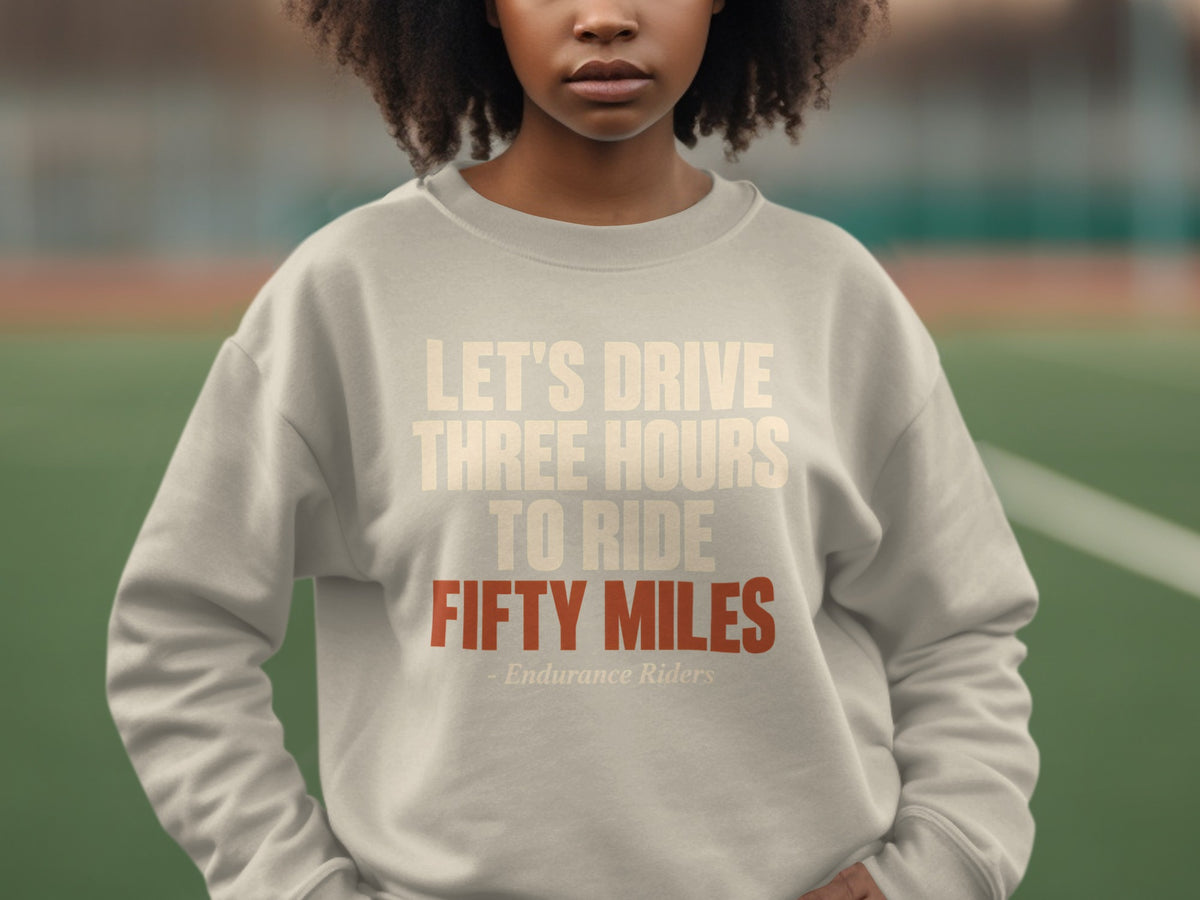 A person wearing a light gray crewneck sweatshirt with the text "LET'S DRIVE THREE HOURS TO RIDE FIFTY MILES - Endurance Riders" printed on the chest.
