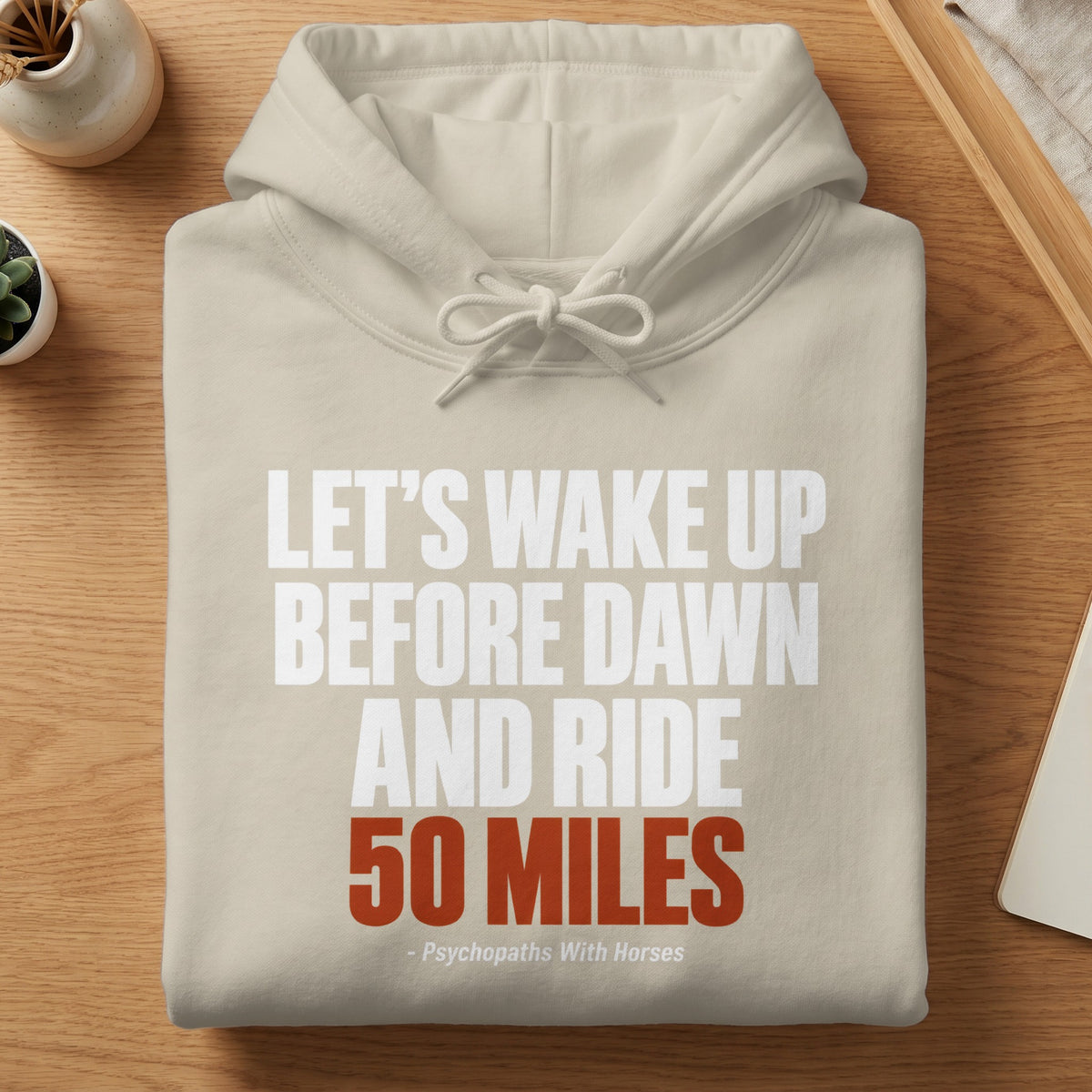 A neatly folded cream-colored hooded sweatshirt lies on a wooden surface, featuring text that reads LET'S WAKE UP BEFORE DAWN AND RIDE 50 MILES.