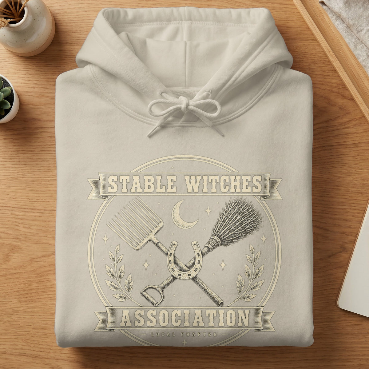 Light grey hoodie with "Stable Witches Association" text and a horseshoe, broom, and pitchfork graphic.