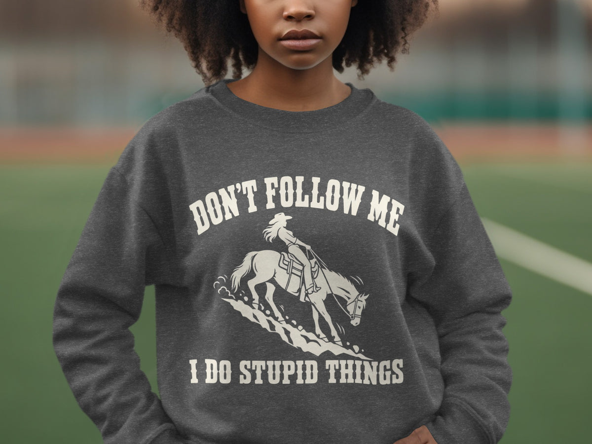 A person wearing a dark grey sweatshirt with white text reading "DON'T FOLLOW ME I DO STUPID THINGS" and a graphic of a cowgirl riding a horse.