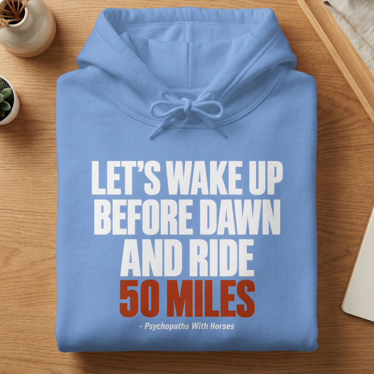 Folded light blue hoodie with white and red text reading Let's wake up before dawn and ride 50 miles - Psychopaths With Horses.