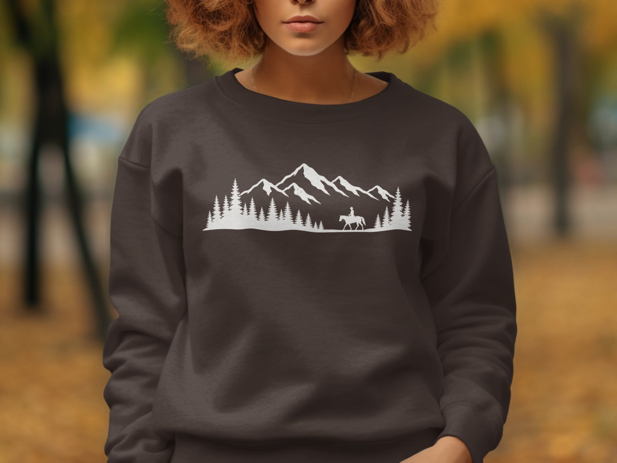 A person wearing a dark brown crewneck sweatshirt featuring a white graphic of mountains, pine trees, and a horseback rider.