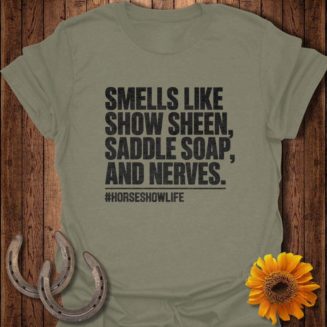 An olive green t-shirt with black text reading "Smells like show sheen, saddle soap, and nerves." and "#horseshowlife".