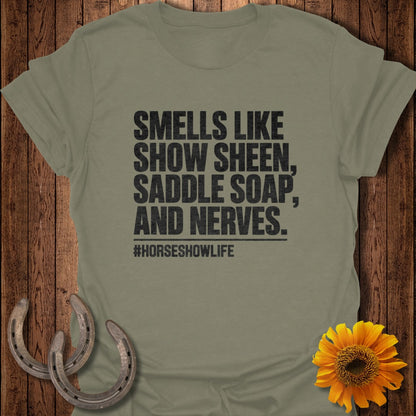 An olive green t-shirt with black text reading "Smells like show sheen, saddle soap, and nerves." and "#horseshowlife".