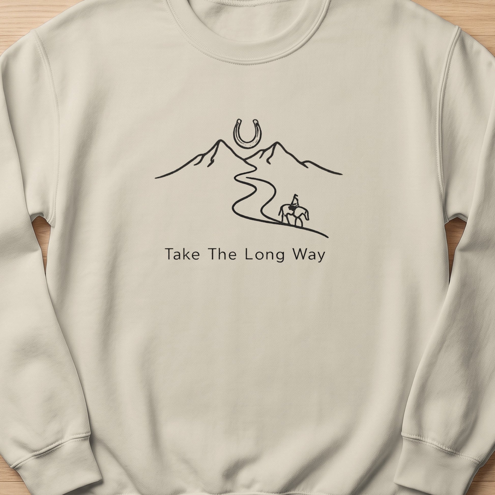 Light cream sweatshirt with black line art of mountains, horseshoe, rider on horseback, and text.