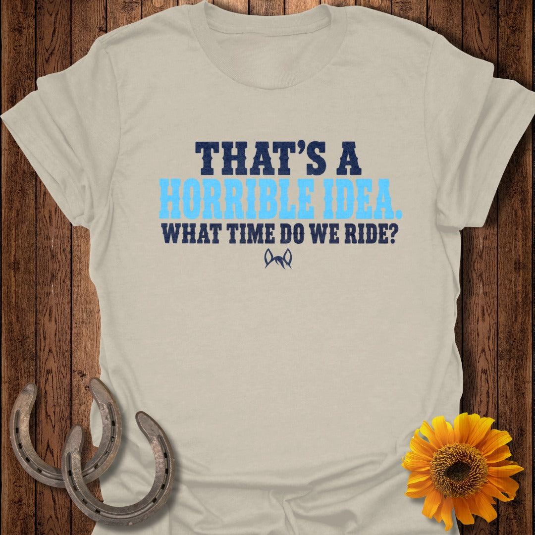 A cream-colored t-shirt with blue text reading That's A Horrible Idea. What Time Do We Ride? on a wooden background.