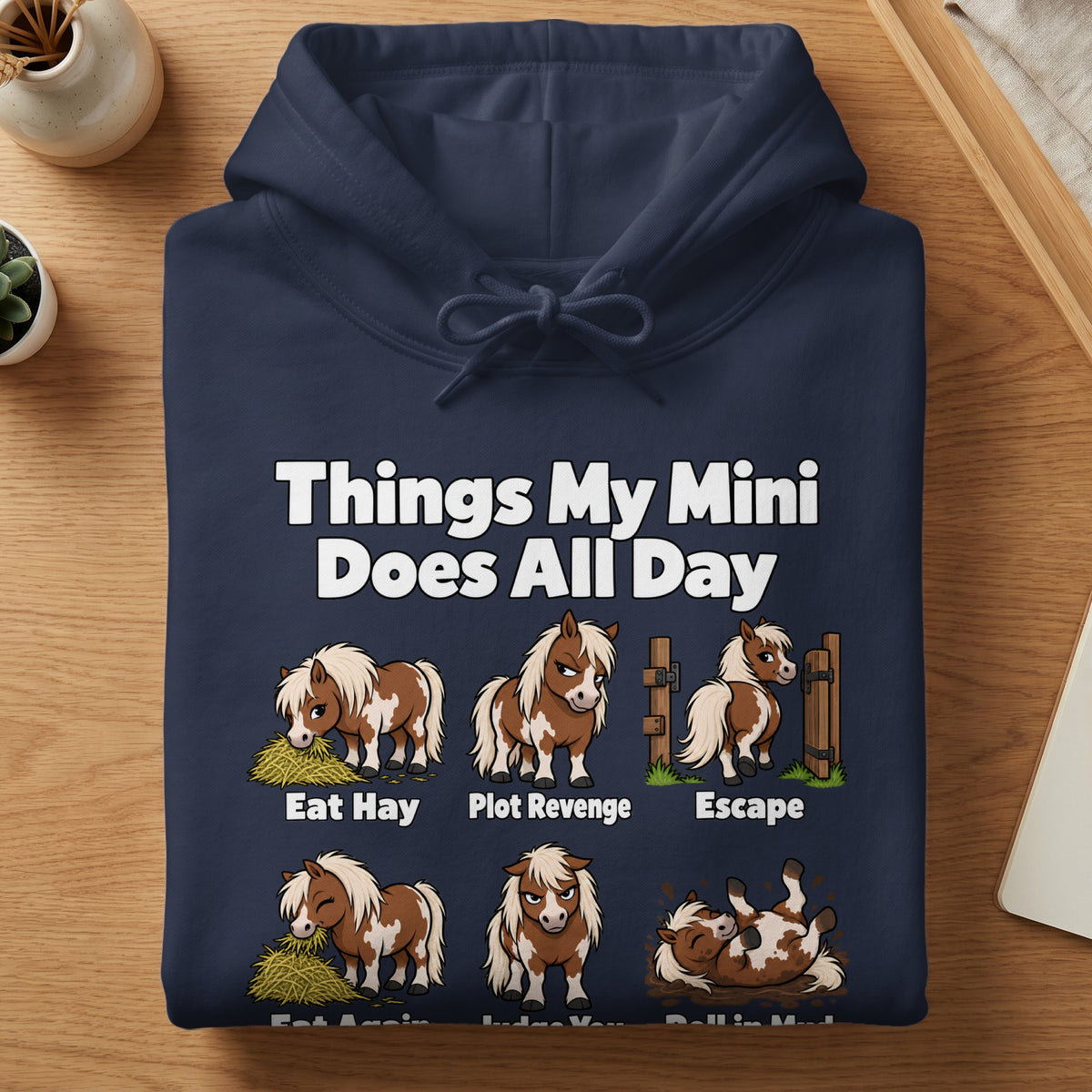 Navy blue hoodie featuring funny illustrations and text about miniature horse behaviors on a wooden table.