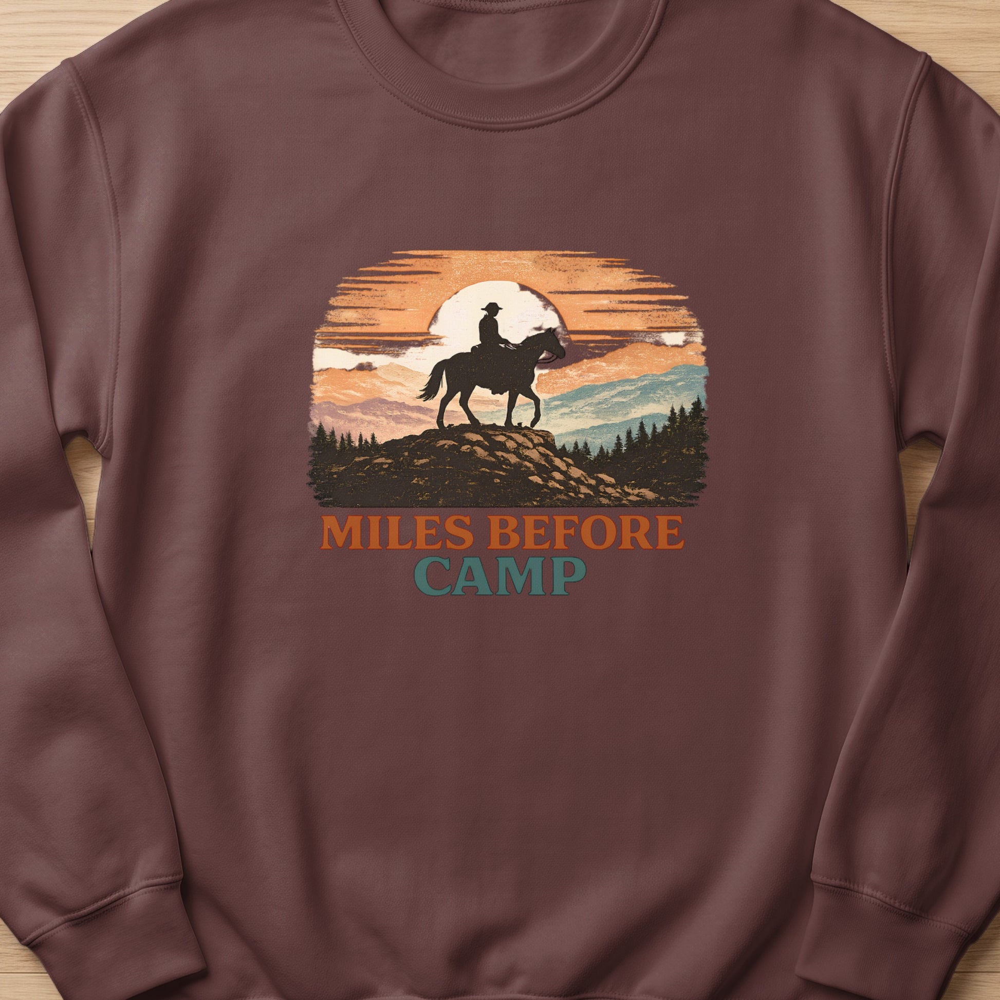 Crewneck sweatshirt with a sunset graphic of a cowboy on horseback with text "MILES BEFORE CAMP".