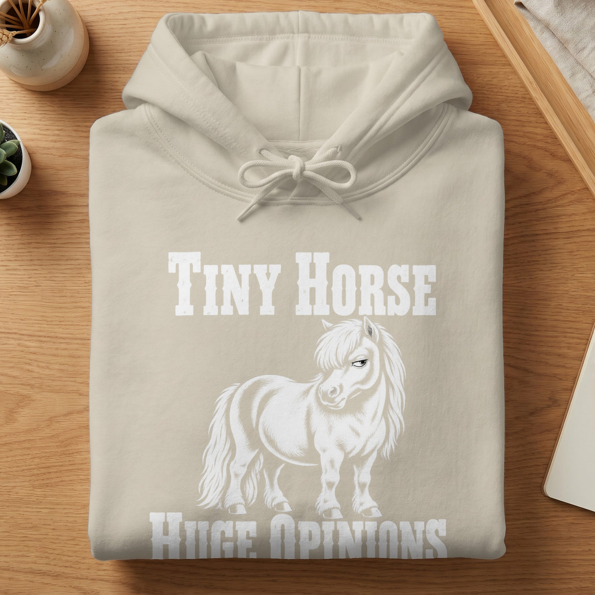 Folded cream-colored hoodie with a white graphic of a pony and the text Tiny Horse Huge Opinions.