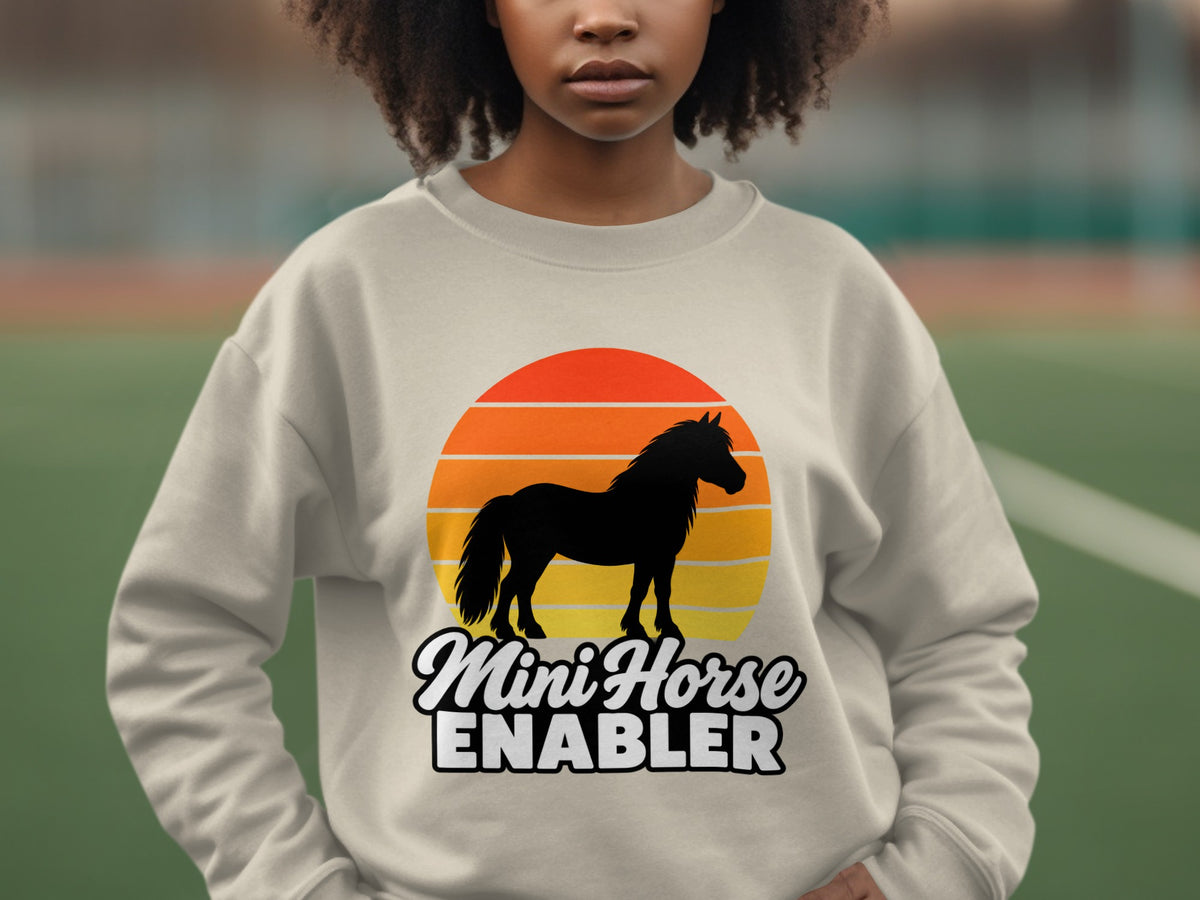 A person wearing a beige crewneck sweatshirt featuring a graphic of a horse and sunset with text reading Mini Horse Enabler.