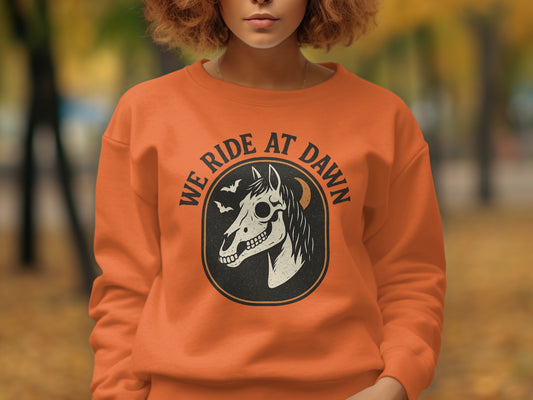 Orange sweatshirt with a circular graphic of a horse skull and bats, text reads "WE RIDE AT DAWN."