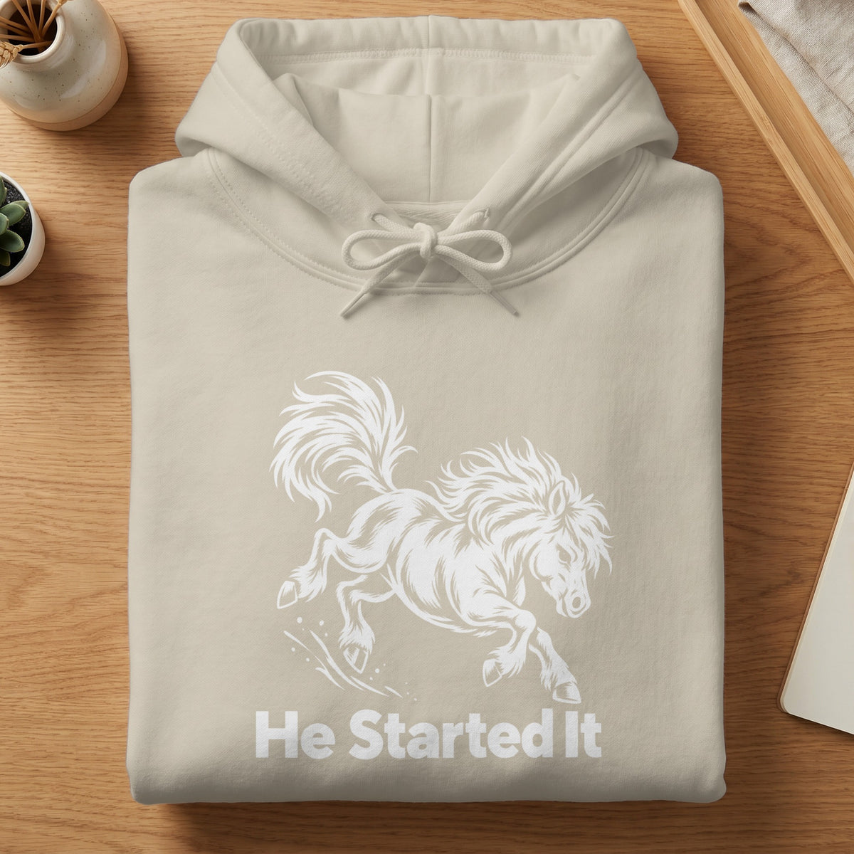 A folded beige hoodie with a white graphic of a running horse and the text He Started It.