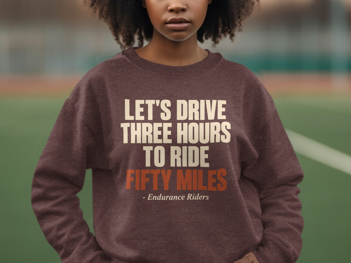 A person wearing a brown sweatshirt with cream and orange text reading, LET'S DRIVE THREE HOURS TO RIDE FIFTY MILES - Endurance Riders.