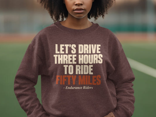 A person wearing a brown sweatshirt with cream and orange text reading, LET'S DRIVE THREE HOURS TO RIDE FIFTY MILES - Endurance Riders.