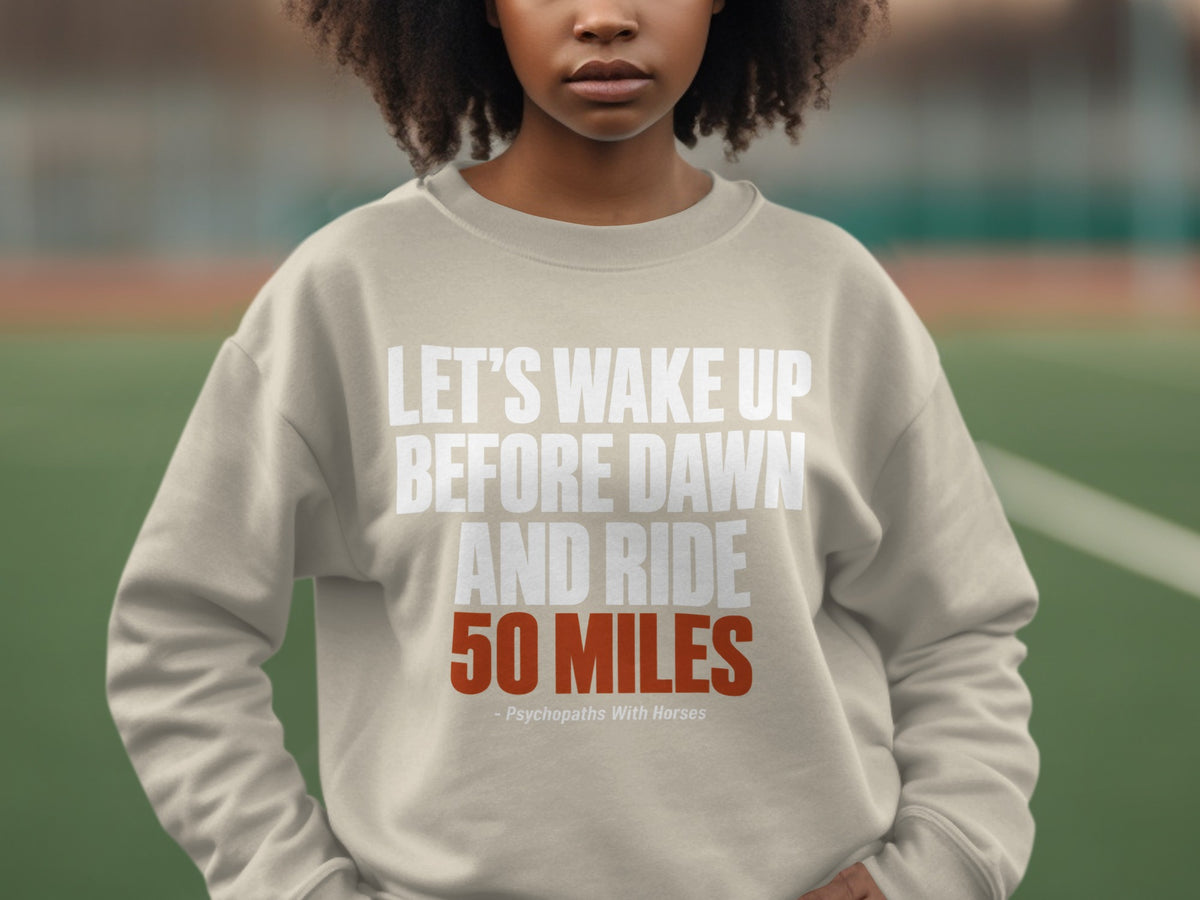 A person wearing a beige crewneck sweatshirt printed with white and orange text that reads Let us wake up before dawn and ride 50 miles.