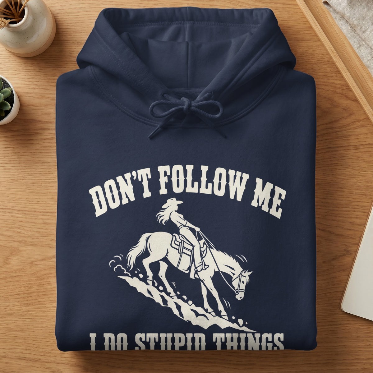 A folded navy blue hoodie featuring white text reading "DON'T FOLLOW ME I DO STUPID THINGS" and a graphic of a cowgirl riding a horse.