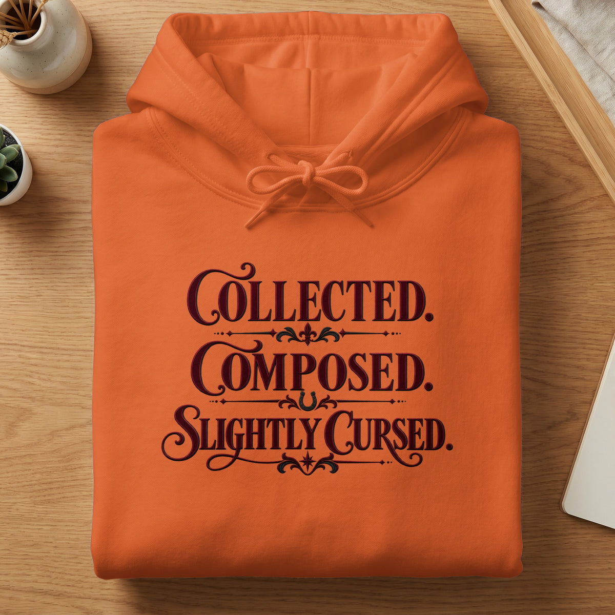 Orange hoodie with "Collected. Composed. Slightly Cursed." text in dark red.