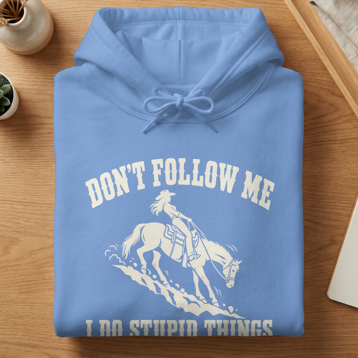 A folded light blue hoodie with white text reading "Don't Follow Me I Do Stupid Things" and a graphic of a cowgirl riding a horse.