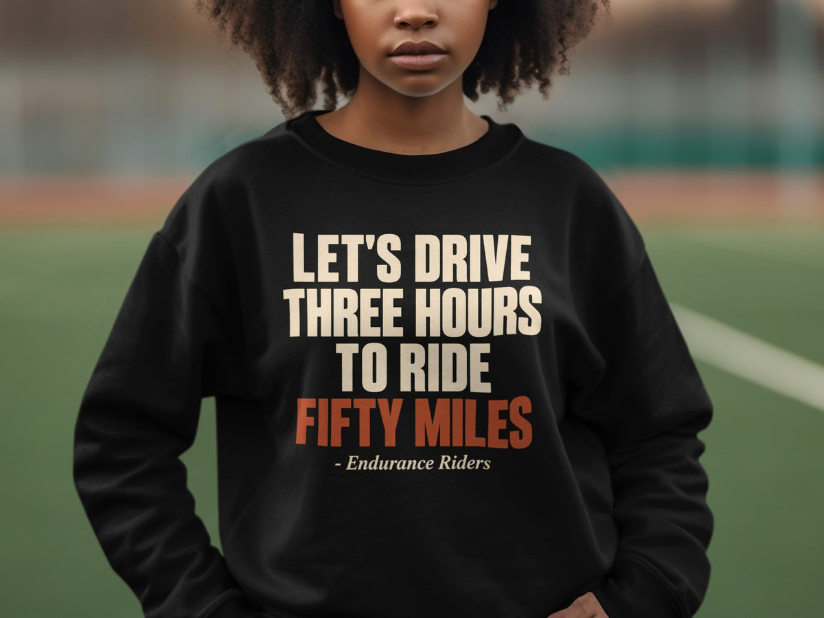 A person with curly hair wears a black sweatshirt featuring the text Let's Drive Three Hours to Ride Fifty Miles - Endurance Riders.
