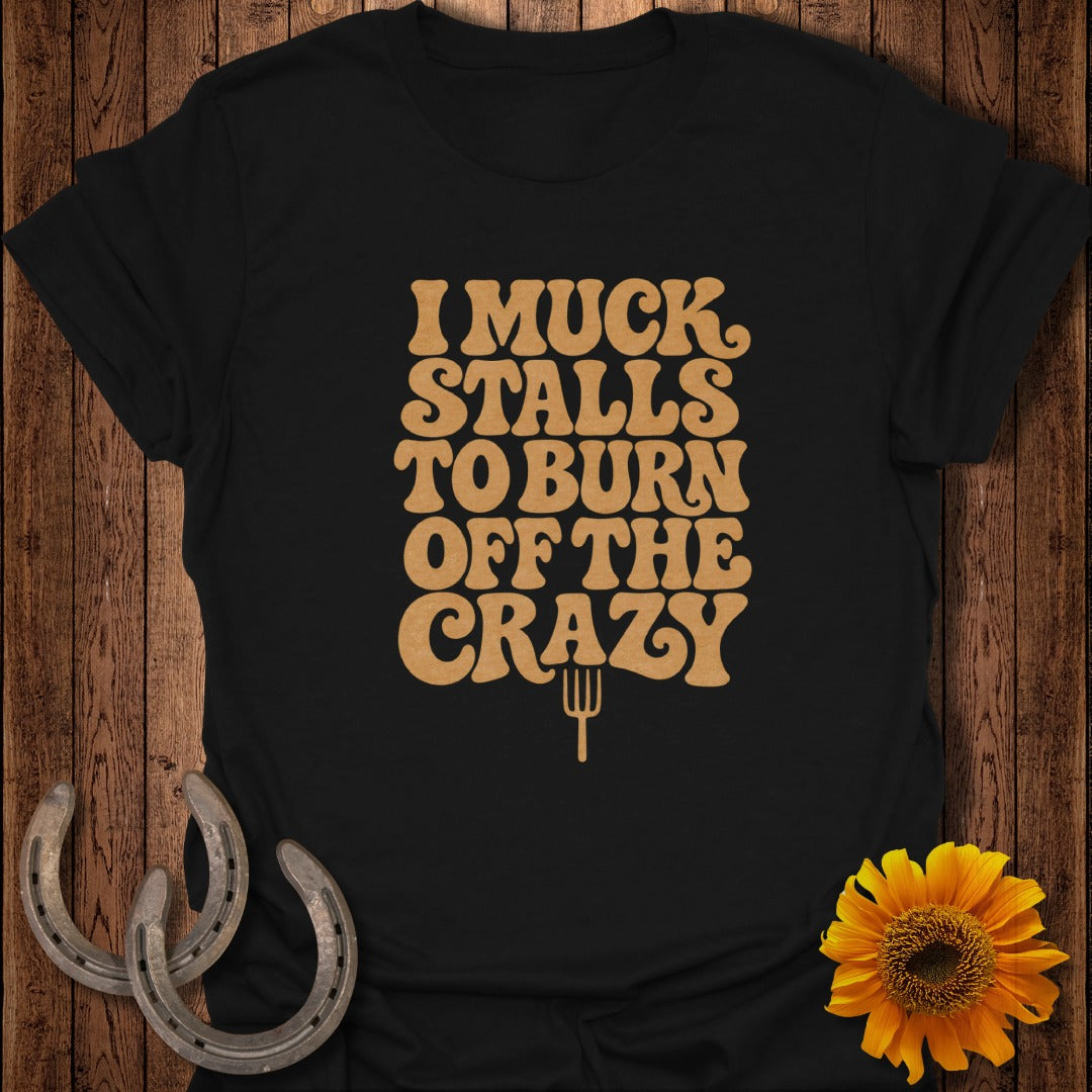 A black t-shirt printed with the tan text "I MUCK STALLS TO BURN OFF THE CRAZY" and a pitchfork icon, laid flat on a wooden background with horseshoes and a sunflower.