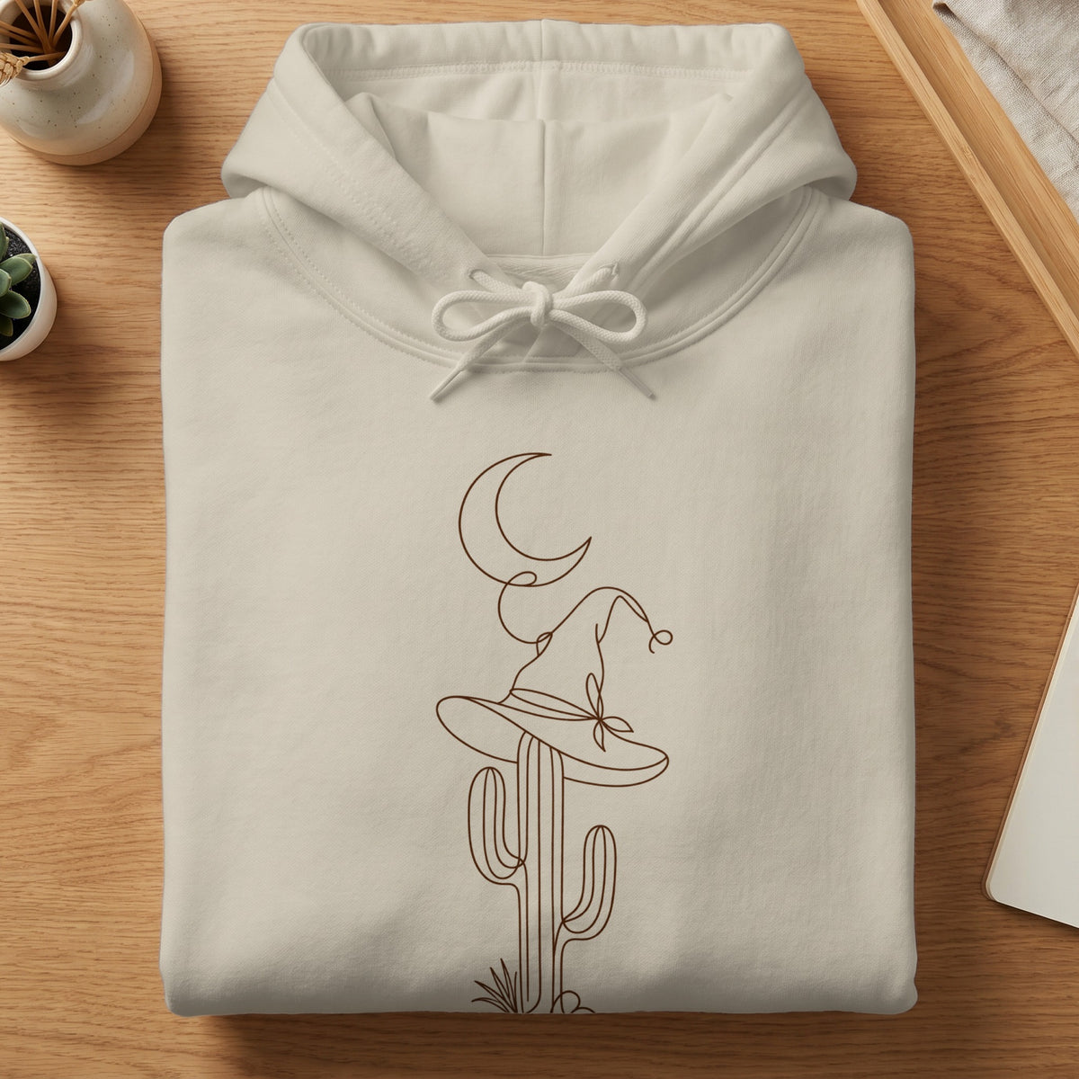 Light beige hoodie with brown line art of a cactus wearing a witch's hat and a crescent moon.