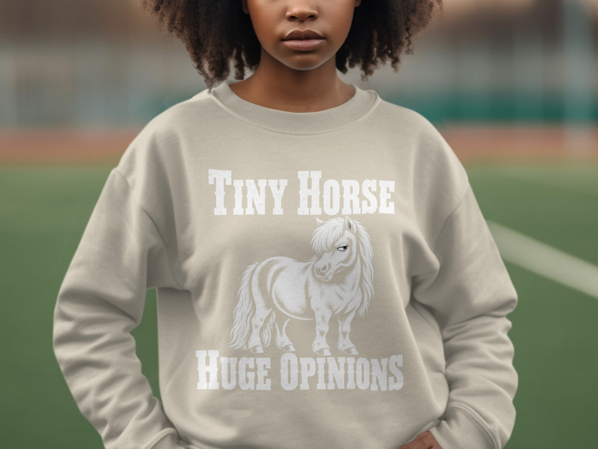 A person wearing a light gray crewneck sweatshirt featuring a graphic of a white pony with the text Tiny Horse Huge Opinions.