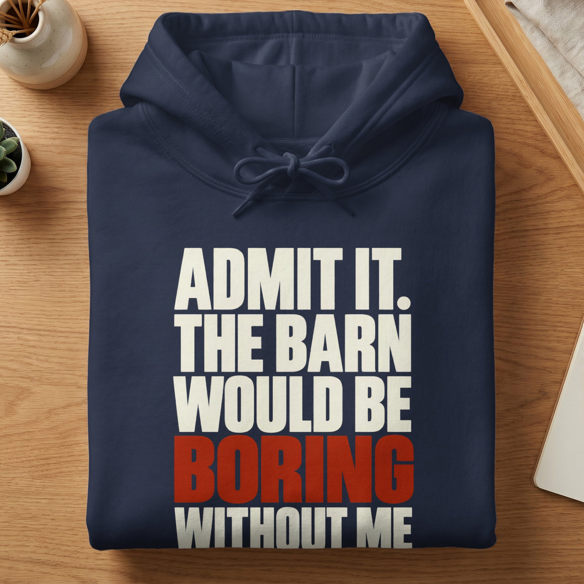 A neatly folded dark blue hoodie on a wooden table, featuring white and red text reading ADMIT IT. THE BARN WOULD BE BORING WITHOUT ME.