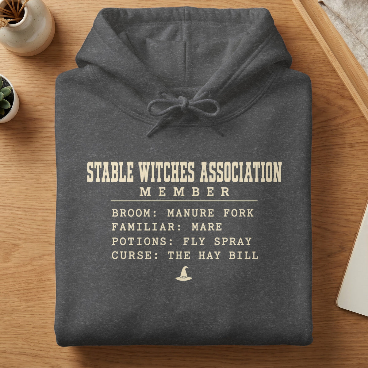 Dark grey hoodie with text: Stable Witches Association Member, Broom, Familiar, Potions, Curse.