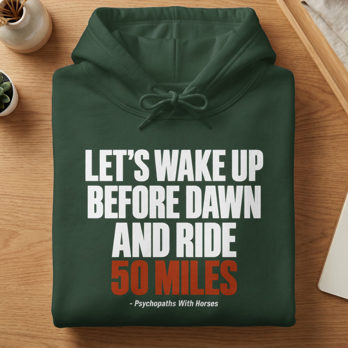 A folded dark green hooded sweatshirt with white and orange text reading Let's wake up before dawn and ride 50 miles - Psychopaths With Horses.