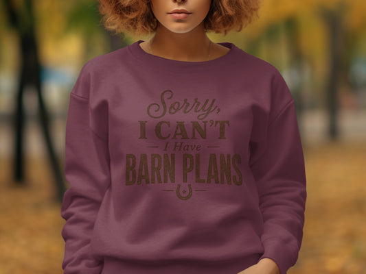 A person wears a maroon sweatshirt printed with the text Sorry, I CAN'T I Have BARN PLANS.
