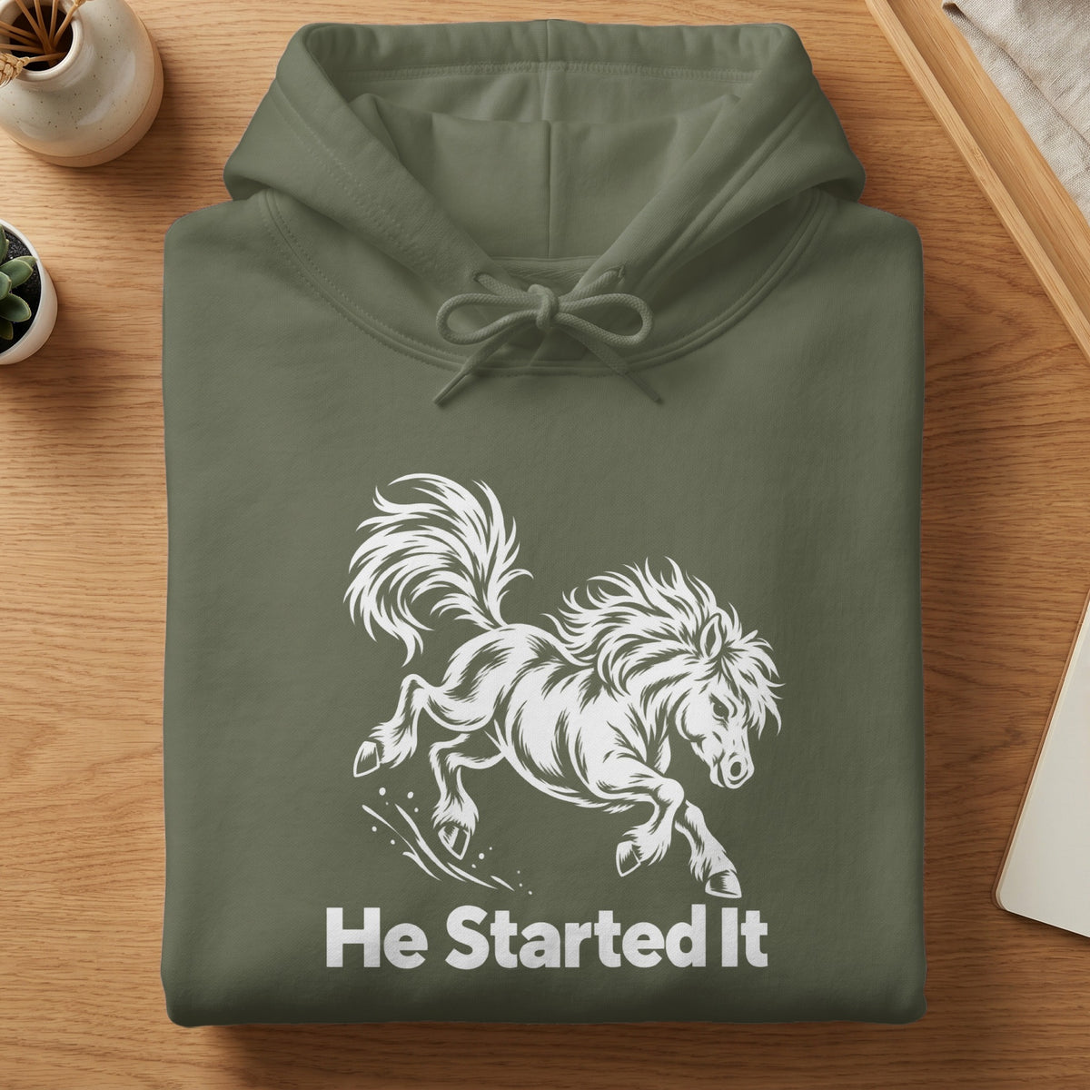 Folded olive green hoodie with a white graphic of a running horse and the text "He Started It".