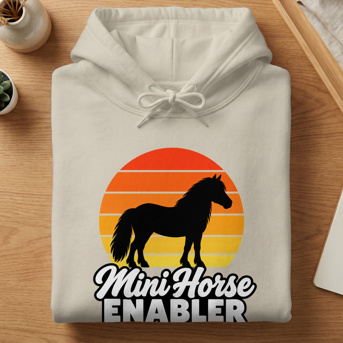 A cream-colored hoodie folded flat on a wooden surface displays a graphic with a black horse silhouette over a retro sunset and the text Mini Horse ENABLER.