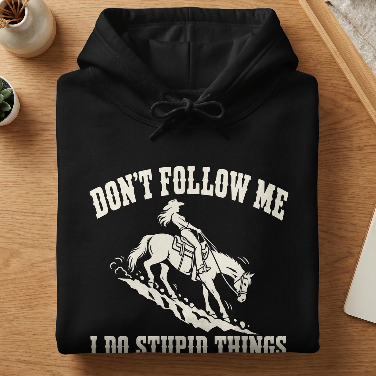 A folded black hoodie features a white graphic of a cowgirl riding a horse with the text "DON'T FOLLOW ME I DO STUPID THINGS."