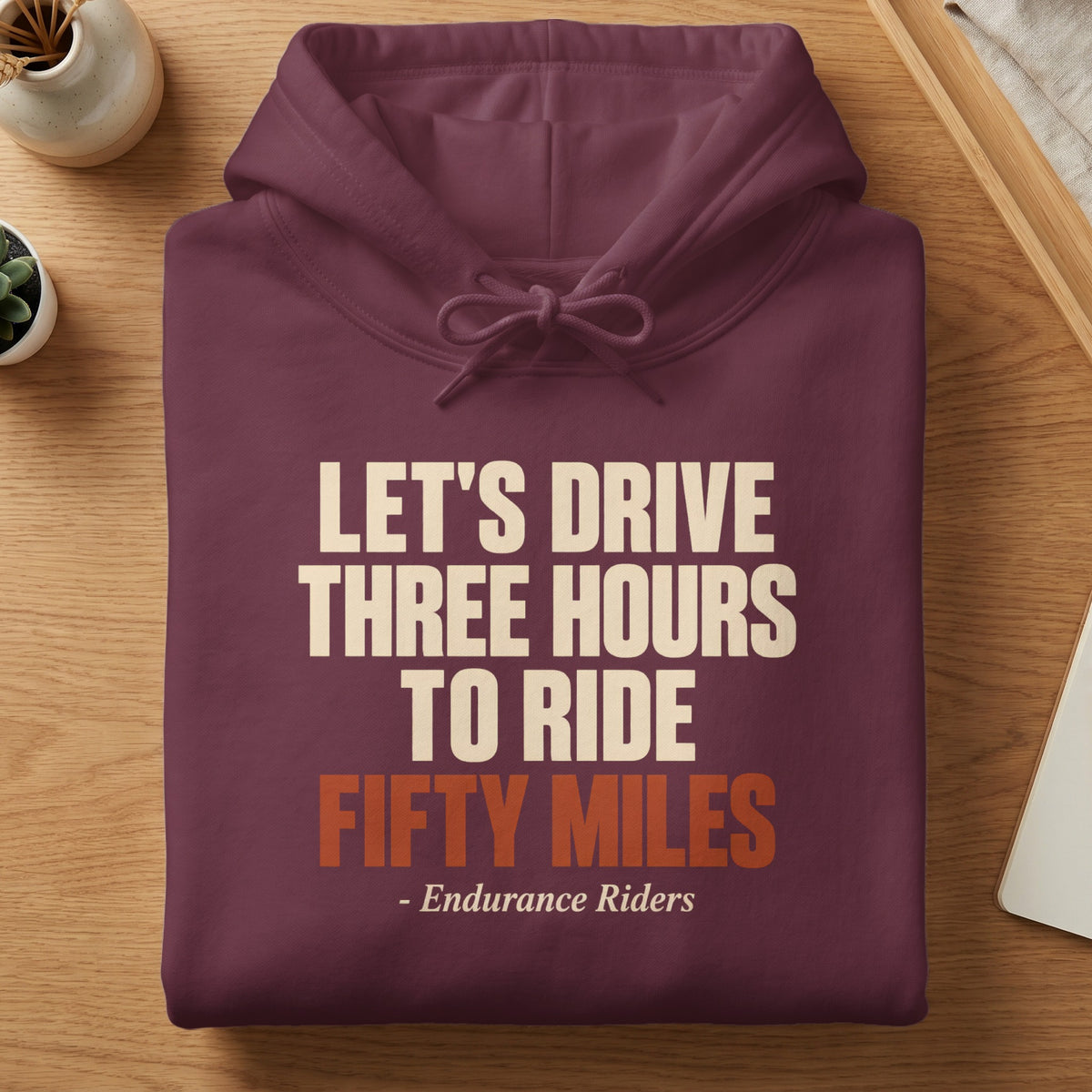 Folded maroon hoodie featuring the cream and orange text, Let's Drive Three Hours To Ride Fifty Miles - Endurance Riders.