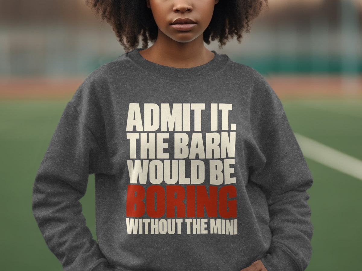 A person wears a dark gray crewneck sweatshirt printed with white and red text stating, Admit it, the barn would be boring without the mini.