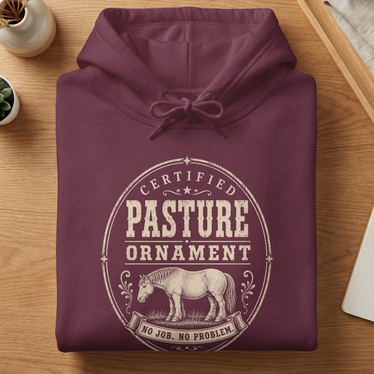 A folded maroon hooded sweatshirt featuring a vintage-style graphic with text reading Certified Pasture Ornament and No Job. No Problem.