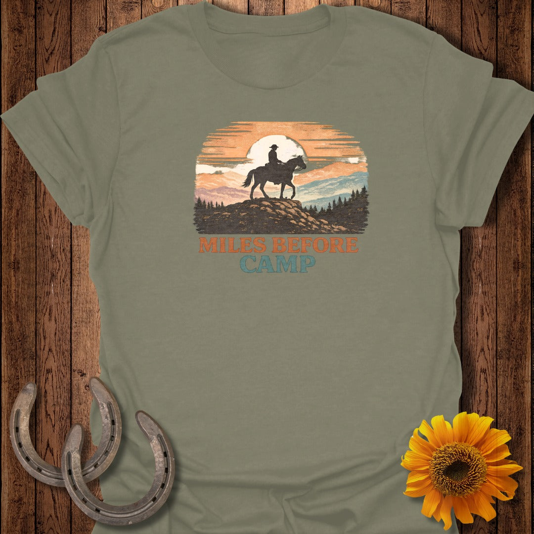Green t-shirt with graphic of a rider on a horse at sunset with text "Miles Before Camp".